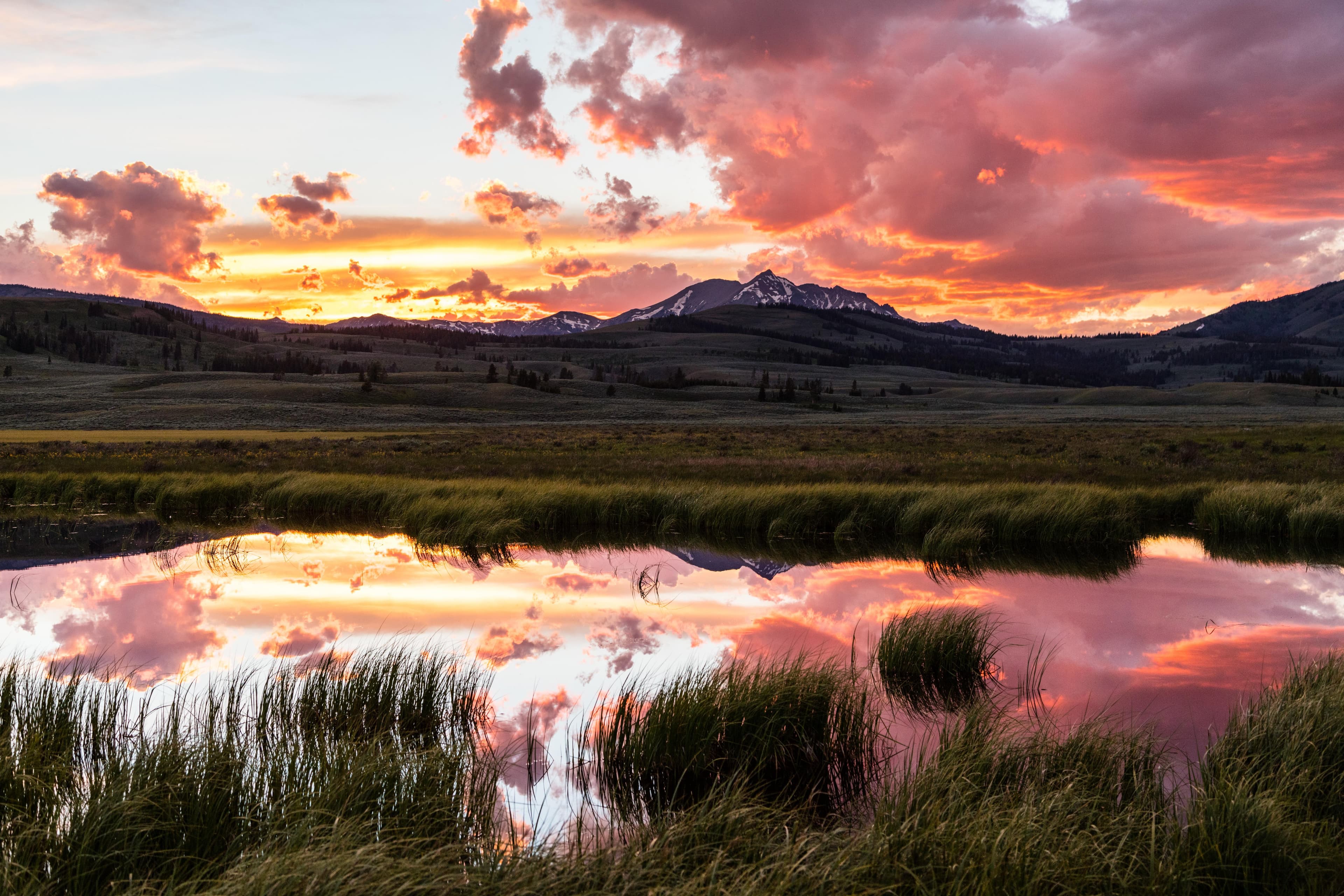 The sun sets behind the sharp point of a snow-covered mountain; pink clouds are reflected in a pond